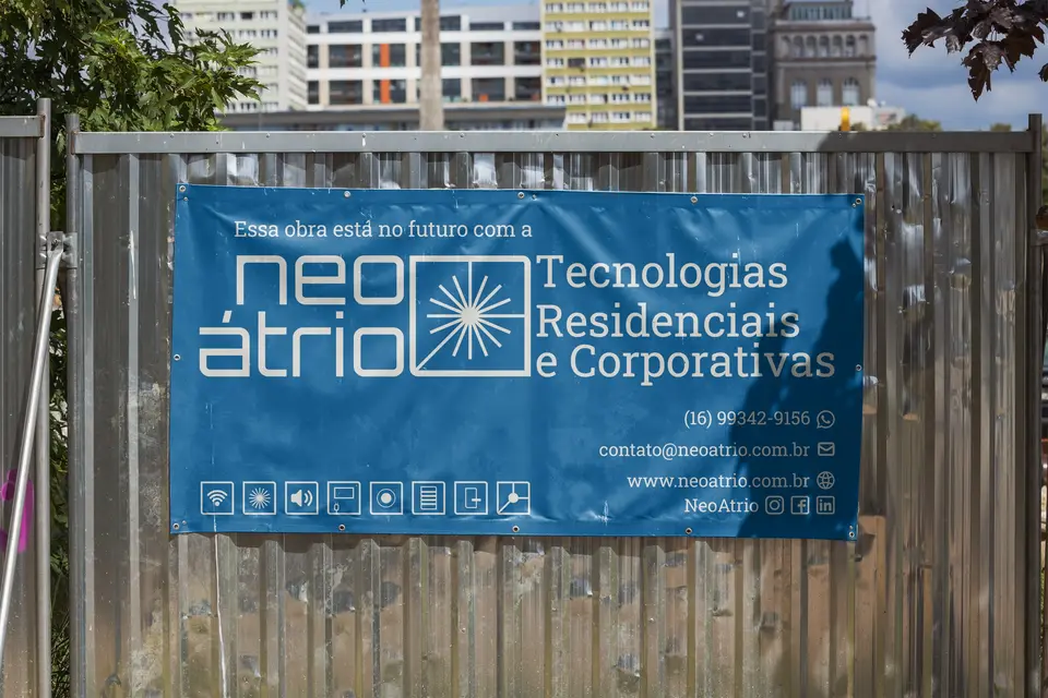Faixa da Neo Átrio presa a um tapume, com o símbolo de raios, o nome e a tagline “Tecnologias residenciais e corporativas”.